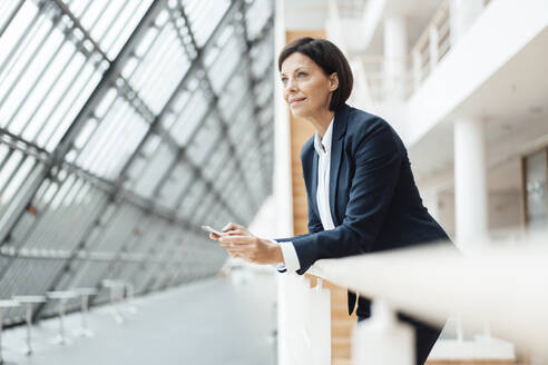 Female professional with smart phone leaning on railing in corridor - JOSEF03782