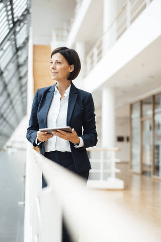 Thoughtful businesswoman with digital tablet looking away by railing in corridor stock photo