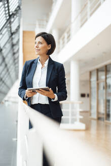 Thoughtful businesswoman with digital tablet looking away by railing in corridor - JOSEF03783