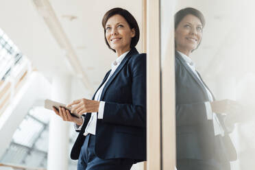 Smiling businesswoman with digital tablet leaning by wall in corridor - JOSEF03785