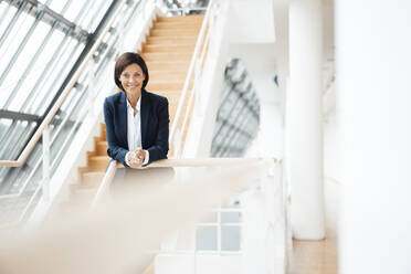 Smiling female professional leaning on railing at corridor - JOSEF03788