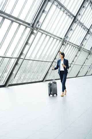 Weibliche Unternehmerin mit Koffer schaut weg, während sie durch einen Korridor geht, lizenzfreies Stockfoto