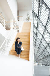 Businesswoman with digital tablet and hand on chin sitting over steps in corridor - JOSEF03808