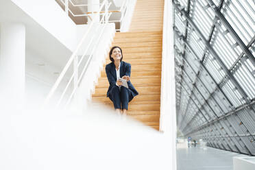 Geschäftsfrau mit digitalem Tablet, die lachend auf einer Treppe im Korridor sitzt - JOSEF03809