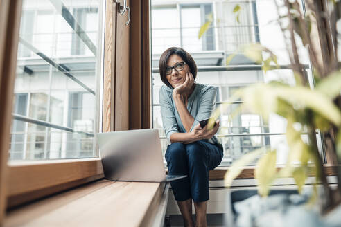 Female entrepreneur with smart phone looking away while sitting in office - JOSEF03813