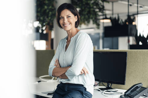 Smiling female entrepreneur with arms crossed against computer in office - JOSEF03820