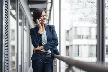 Happy businesswoman talking on mobile phone on office balcony - JOSEF03832