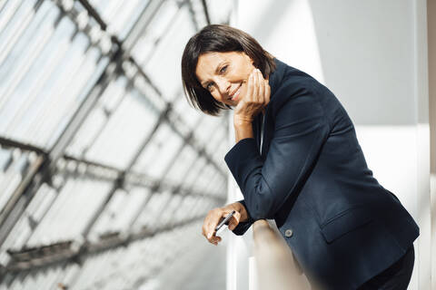 Smiling businesswoman with hand on chin leaning on railing on corridor stock photo