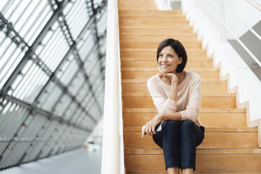 Lächelnde Geschäftsfrau mit Hand am Kinn auf einer Treppe im Korridor sitzend - JOSEF03838