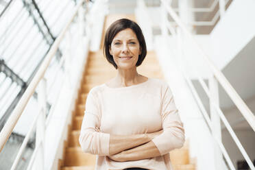 Smiling confident businesswoman with arms crossed against steps in corridor - JOSEF03840