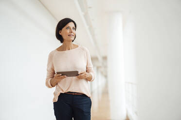 Reife weibliche Fachkraft mit digitalem Tablet, die wegschaut, während sie auf einem Korridor steht - JOSEF03846