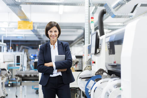 Confident businesswoman with digital tablet standing by machinery in factory - JOSEF03856