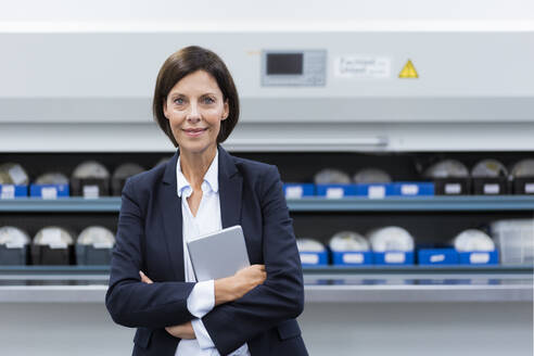 Smiling female entrepreneur with arms crossed and digital tablet at industry - JOSEF03861