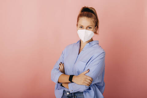 Businesswoman wearing FFP2 face mask standing with arms crossed against colored background stock photo