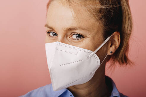 Businesswoman wearing protective face mask staring while standing against colored background - DAWF01785