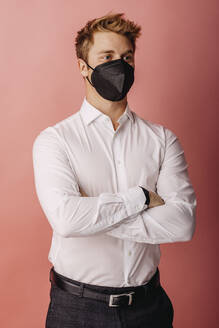 Männlicher Unternehmer mit FFP2-Gesichtsmaske, der wegschaut, während er mit verschränkten Armen vor einem farbigen Hintergrund steht - DAWF01794