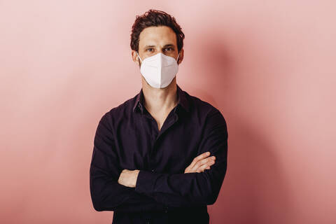 Mid adult businessman wearing FFP2 face mask standing with arms crossed against colored background stock photo