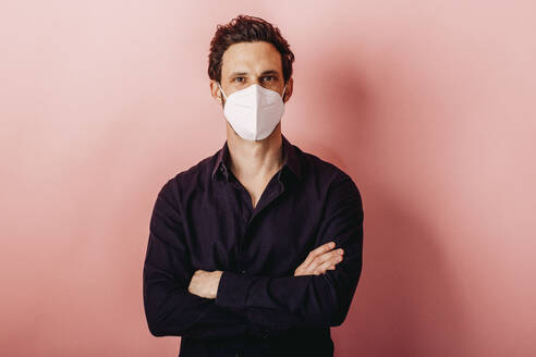 Mid adult businessman wearing FFP2 face mask standing with arms crossed against colored background - DAWF01796