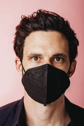 Businessman wearing black color protective face mask staring while standing against colored background - DAWF01801