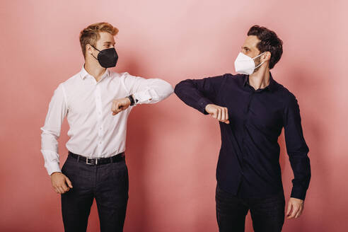 Männlicher Unternehmer mit Schutzmaske grüßt mit Ellbogenstößen, während er vor einem farbigen Hintergrund steht - DAWF01810