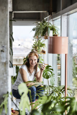 Smiling mature woman with headphones listening music while having coffee in loft apartment stock photo