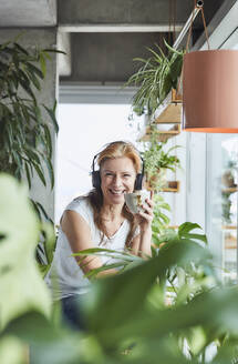 Glückliche Frau, die eine Kaffeetasse hält und über Kopfhörer Musik hört, während sie in einer Loftwohnung sitzt - FMKF06975