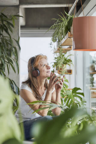 Lächelnde Frau mit Kopfhörern, die eine Kaffeetasse hält und dabei wegschaut, in einer Loftwohnung zu Hause, lizenzfreies Stockfoto