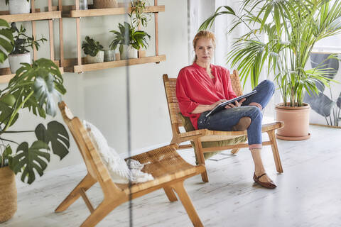 Reife Frau in Freizeitkleidung mit Laptop sitzt auf Stuhl in Loft-Wohnung zu Hause, lizenzfreies Stockfoto