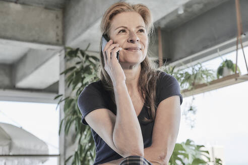 Lächelnde Frau, die wegschaut, während sie in einer Dachgeschosswohnung zu Hause mit dem Handy telefoniert - FMKF06980