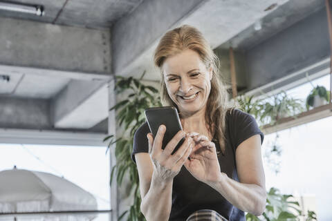 Lächelnde reife Frau, die ein Mobiltelefon benutzt, während sie zu Hause in einer Loftwohnung sitzt, lizenzfreies Stockfoto