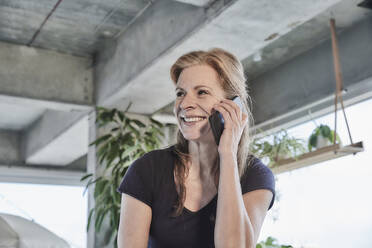 Glückliche reife Frau, die wegschaut, während sie mit einem Smartphone in einer Loftwohnung zu Hause spricht - FMKF06982