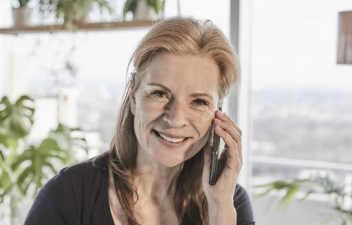Schöne lächelnde Frau mit braunen Haaren im Gespräch auf dem Handy in Loft-Wohnung zu Hause - FMKF06983
