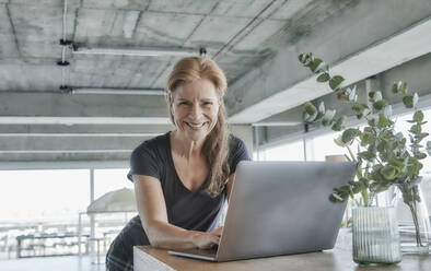 Lächelnde reife Frau mit Laptop am Tisch in Loft-Wohnung zu Hause - FMKF06985
