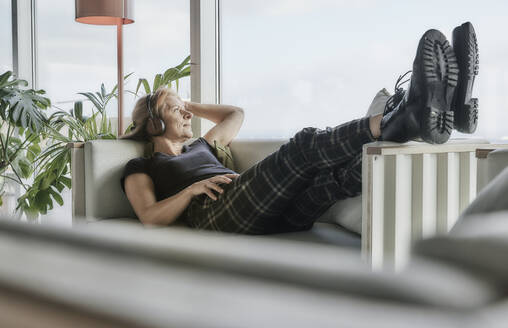Reife Frau mit hochgelegten Füßen hört Musik über Kopfhörer, während sie zu Hause auf dem Sofa liegt - FMKF06988
