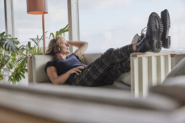 Woman with hand in hair listening music while lying on sofa at home - FMKF06989