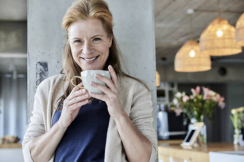 Lächelnde reife Frau hält Kaffeetasse gegen Säule zu Hause, lizenzfreies Stockfoto