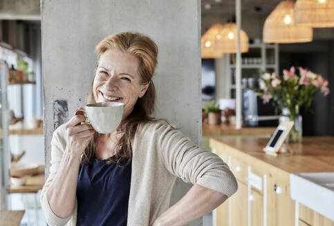 Glückliche reife Frau mit Hand auf der Hüfte beim Kaffee trinken gegen Säule zu Hause - FMKF06995