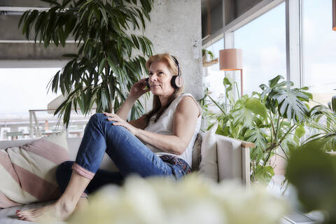 Frau mit Kopfhörern hört Musik, während sie auf dem Sofa in der Wohnung sitzt, lizenzfreies Stockfoto