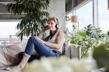 Woman with headphones listening music while sitting on sofa at apartment - FMKF06996