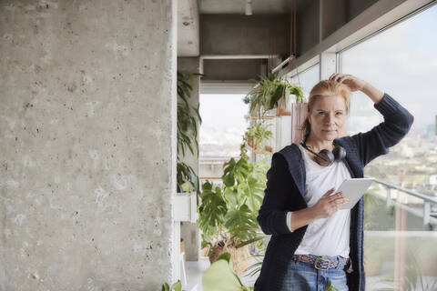 Frau mit Hand in den Haaren, die ein digitales Tablet hält, während sie sich auf ein Glas in einer Loftwohnung zu Hause stützt, lizenzfreies Stockfoto