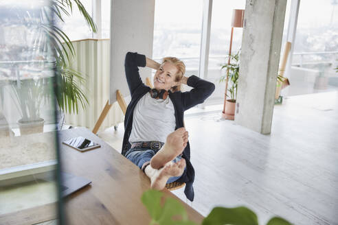 Smiling businesswoman looking away while sitting with feet up on desk at home - FMKF07008