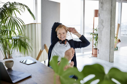 Lächelnde Unternehmerin mit Händen hinter dem Kopf, die sich auf einem Stuhl am Schreibtisch im Heimbüro entspannt - FMKF07009