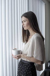 Junge Frau mit Kaffeetasse, die wegschaut, während sie zu Hause am Fenster steht - AJOF01091