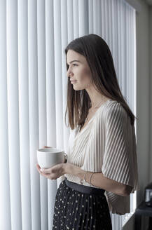 Young woman with coffee cup looking away while standing by window at home - AJOF01091
