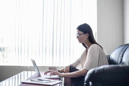 Junge Frau mit Brille, die zu Hause sitzend am Laptop arbeitet - AJOF01092