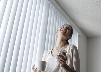 Lächelnde Frau mit Kaffeetasse, die ein Mobiltelefon benutzt, während sie zu Hause am Fenster steht - AJOF01095