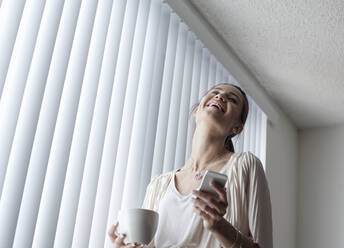 Smiling woman with coffee cup using mobile phone while standing by window at home - AJOF01095