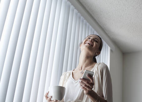 Lächelnde Frau mit Kaffeetasse, die ein Mobiltelefon benutzt, während sie zu Hause am Fenster steht - AJOF01095