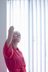 Woman stretching hands while standing by window at home - AJOF01097