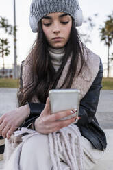 Teenager-Mädchen benutzt Mobiltelefon, während sie auf einer Parkbank sitzt - JRVF00284